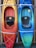 Two kayaks are mounted vertically on a wooden rack. The kayak on the left has a gradient from yellow to orange, while the one on the right transitions from blue to green. Both kayaks are equipped with cushioned seats and elastic straps for securing gear.