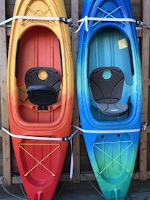 Two kayaks are mounted vertically on a wooden rack. The kayak on the left has a gradient from yellow to orange, while the one on the right transitions from blue to green. Both kayaks are equipped with cushioned seats and elastic straps for securing gear.
