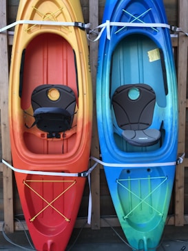 Two kayaks are mounted vertically on a wooden rack. The kayak on the left has a gradient from yellow to orange, while the one on the right transitions from blue to green. Both kayaks are equipped with cushioned seats and elastic straps for securing gear.