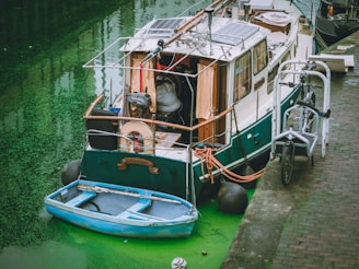 A small, eclectic boat docked along a canal with murky green water. The boat has solar panels on its roof, various ropes and equipment on deck, and a smaller blue dinghy tied to it. Bicycles are parked on the nearby brick pavement.