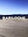 Large white letters spelling 'BARILOCHE' are placed on a cement surface with a lake and mountains in the background. The sky is clear and blue, indicating a sunny day.