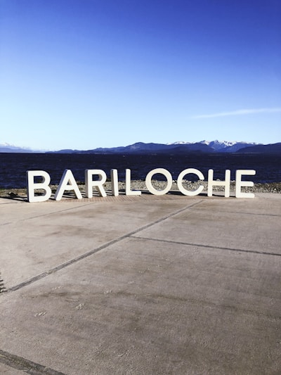 Large white letters spelling 'BARILOCHE' are placed on a cement surface with a lake and mountains in the background. The sky is clear and blue, indicating a sunny day.