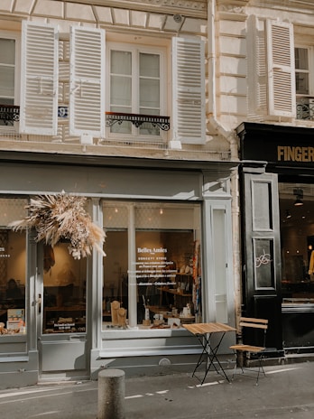 The image depicts a charming storefront with the name 'Belles Amies' on the window. The store has a rustic and inviting exterior, featuring a display of items through large glass windows. A dried flower wreath decorates the entrance. Outside the shop, there is a small, wooden, foldable table with two chairs. The building has classic architectural elements, including shutters on the windows above the store.