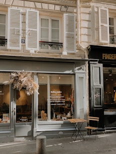 The image depicts a charming storefront with the name 'Belles Amies' on the window. The store has a rustic and inviting exterior, featuring a display of items through large glass windows. A dried flower wreath decorates the entrance. Outside the shop, there is a small, wooden, foldable table with two chairs. The building has classic architectural elements, including shutters on the windows above the store.