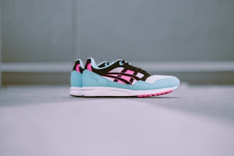 A pair of stylish athletic sneakers is placed on a flat surface. The sneakers feature a striking color combination of pastel blue, black, white, and vibrant pink. The design includes a sleek logo on the side and a textured sole.