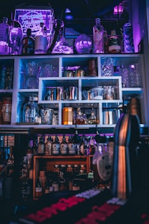 A bar setup with various bottles of liquor and syrups, arranged on shelves. Glassware and cocktail shakers are neatly organized, and a neon sign in the background casts a purple hue, enhancing the ambience. The lighting is dim, with accents of blue and pink, creating a moody effect.