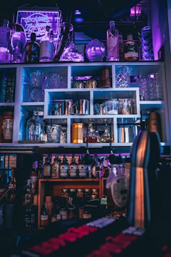 A bar setup with various bottles of liquor and syrups, arranged on shelves. Glassware and cocktail shakers are neatly organized, and a neon sign in the background casts a purple hue, enhancing the ambience. The lighting is dim, with accents of blue and pink, creating a moody effect.