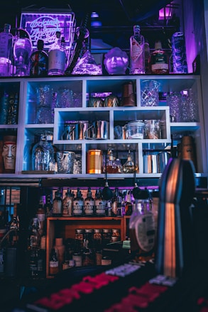 A bar setup with various bottles of liquor and syrups, arranged on shelves. Glassware and cocktail shakers are neatly organized, and a neon sign in the background casts a purple hue, enhancing the ambience. The lighting is dim, with accents of blue and pink, creating a moody effect.