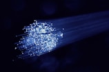 A cluster of glowing fiber optic cables emitting bright blue light against a dark background. The cables appear to be bundled together, with individual fibers visible, creating a starry effect as they disperse light.