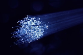 A cluster of glowing fiber optic cables emitting bright blue light against a dark background. The cables appear to be bundled together, with individual fibers visible, creating a starry effect as they disperse light.