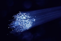 A cluster of glowing fiber optic cables emitting bright blue light against a dark background. The cables appear to be bundled together, with individual fibers visible, creating a starry effect as they disperse light.