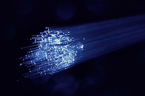 A cluster of glowing fiber optic cables emitting bright blue light against a dark background. The cables appear to be bundled together, with individual fibers visible, creating a starry effect as they disperse light.
