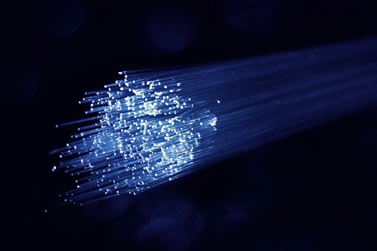 A cluster of glowing fiber optic cables emitting bright blue light against a dark background. The cables appear to be bundled together, with individual fibers visible, creating a starry effect as they disperse light.