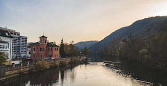 A peaceful river flows through a picturesque landscape, with a small, peach-colored building with a tower by its side, and modern apartments nearby. The scene is surrounded by lush, verdant hills and trees, gently illuminated by a sun setting behind the hills, creating a tranquil atmosphere.