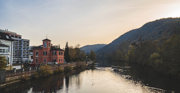 A peaceful river flows through a picturesque landscape, with a small, peach-colored building with a tower by its side, and modern apartments nearby. The scene is surrounded by lush, verdant hills and trees, gently illuminated by a sun setting behind the hills, creating a tranquil atmosphere.