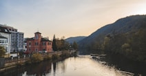 A peaceful river flows through a picturesque landscape, with a small, peach-colored building with a tower by its side, and modern apartments nearby. The scene is surrounded by lush, verdant hills and trees, gently illuminated by a sun setting behind the hills, creating a tranquil atmosphere.