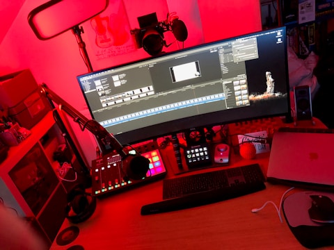 A workspace setup featuring a large curved monitor displaying video editing software. It is surrounded by various equipment including a microphone on an adjustable arm, a sound mixer with illuminated buttons, headphones, and a laptop. The ambient lighting in the room is predominantly red, creating an intense and focused atmosphere.