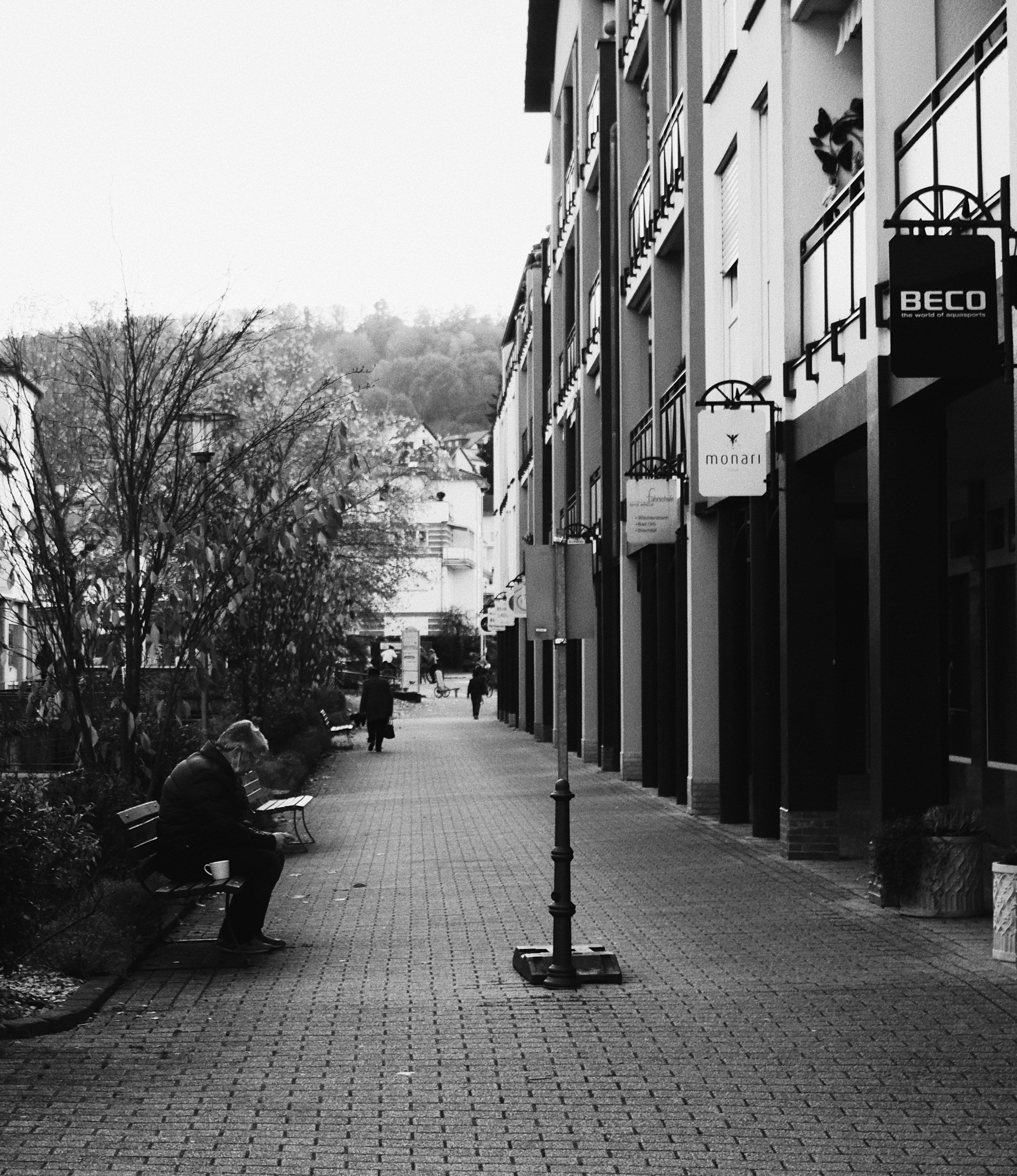 grayscale photo of man sitting on bench near bare trees and buildings