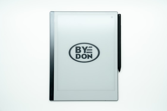 A digital tablet with an e-ink screen displays the phrase 'BYE DON' inside an oval, suggesting it might be a play on words or statement. A stylus is attached on the right side of the device.