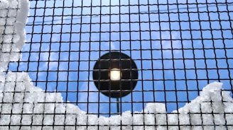 A metal grid is covered with patches of snow, set against a bright, clear blue sky. A circular light fixture is visible through the grid, providing a contrasting focus point.