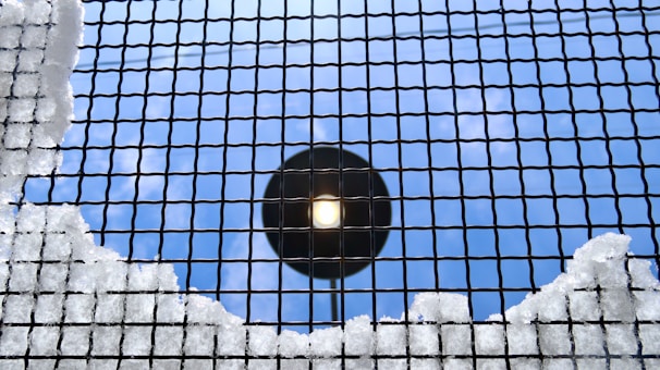 A metal grid is covered with patches of snow, set against a bright, clear blue sky. A circular light fixture is visible through the grid, providing a contrasting focus point.