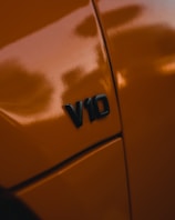 A close-up view of a car with an orange paint job featuring a 'V10' badge, indicating the engine type. The glossy surface reflects light, highlighting the sleek design and vibrant color.
