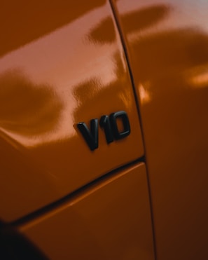 A close-up view of a car with an orange paint job featuring a 'V10' badge, indicating the engine type. The glossy surface reflects light, highlighting the sleek design and vibrant color.