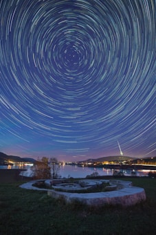 A breathtaking night sky filled with star trails forming circular patterns above a tranquil landscape. The foreground features a stone labyrinth, while a calm lake reflects the lights from distant buildings and hills. A distinct tower with an illuminated spire is visible on the horizon, adding a focal point to the serene scene.