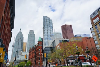 A bustling cityscape featuring a mix of modern skyscrapers and historic brick buildings under a partly cloudy sky. Trees with green foliage line the street, and the CN Tower is visible in the skyline. Traffic lights and signs are present on the busy street.