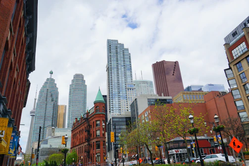A bustling cityscape featuring a mix of modern skyscrapers and historic brick buildings under a partly cloudy sky. Trees with green foliage line the street, and the CN Tower is visible in the skyline. Traffic lights and signs are present on the busy street.