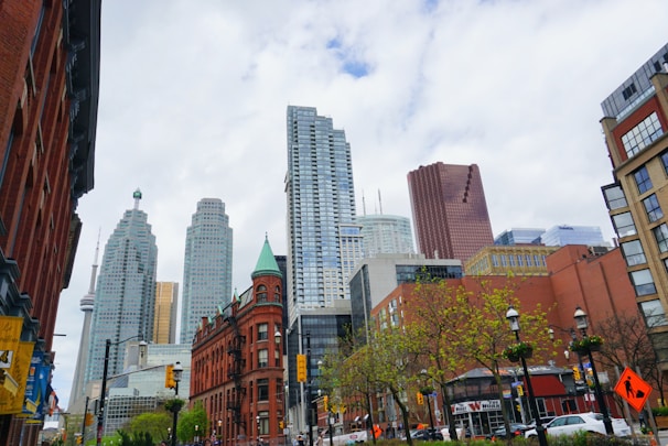 A bustling cityscape featuring a mix of modern skyscrapers and historic brick buildings under a partly cloudy sky. Trees with green foliage line the street, and the CN Tower is visible in the skyline. Traffic lights and signs are present on the busy street.