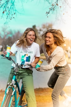 Two people are laughing and enjoying each other's company outdoors. One person is holding a bike and a can, wearing a white t-shirt with a graphic. The other person is holding a similar can, wearing a light sweater and jeans. The scene is bright, with bare tree branches in the background and a lake nearby.