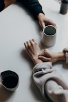 Two hands with cups of coffee are on a table. One hand is gently holding or comforting the wrist of the other. The scene is intimate and suggests a moment of connection or support.
