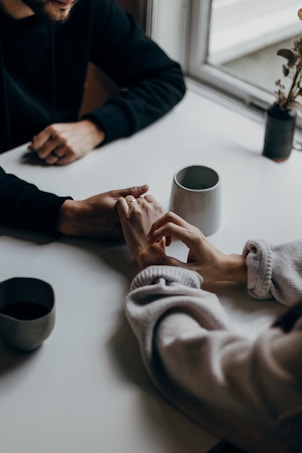 A close-up view of two people sitting at a table, holding hands in a gentle and comforting manner. The table has a few ceramic objects, including two cups and a small vase with dried flowers. Natural light is coming through a nearby window, creating a soft and intimate atmosphere.