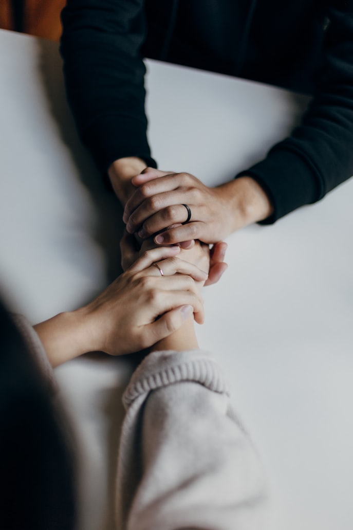 Two people are seen holding hands across a table, with only their arms visible. Both people wear rings on their left ring fingers. One wears a black long-sleeved sweater, and the other a gray one.