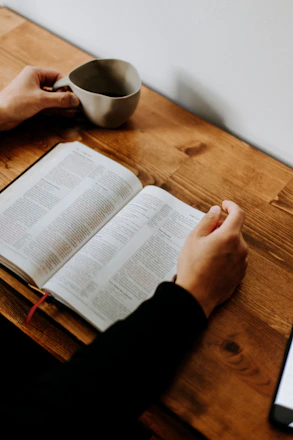 A person reading an open book on a wooden table with a coffee mug