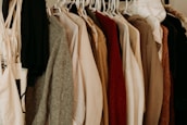 A variety of clothes hung on white plastic hangers, including sweaters, jackets, and shirts, showcasing a mix of textures like wool and cotton in neutral and earthy tones.