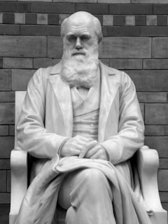 A marble statue of a seated man with a beard, holding a stack of papers on his lap. He appears to be wearing a suit, and his expression is thoughtful. Behind the statue is a brick wall, adding a textured backdrop.