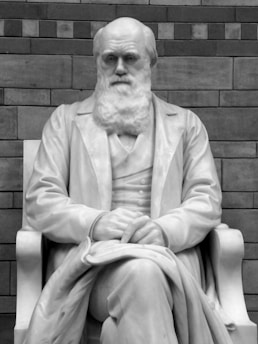 A marble statue of a seated man with a beard, holding a stack of papers on his lap. He appears to be wearing a suit, and his expression is thoughtful. Behind the statue is a brick wall, adding a textured backdrop.