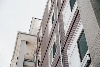 The image features a modern building with a geometric design. The structure has a facade of brown horizontal lines and large white-framed windows. The perspective is skewed slightly upwards, showcasing an angled view of the architecture against a pale, overcast sky.