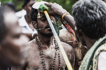 A man is engaged in a religious ritual, with a long metal rod piercing his cheeks. He is adorned with a beaded necklace and has various colorful fabrics and objects placed on his head. Surrounding him are other individuals, focused on the ritual.