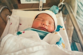 A newborn baby is peacefully sleeping in a hospital bassinet, wrapped in a white blanket with colorful designs. The infant is wearing a small hat, and the setting appears to be a hospital or a nursery.