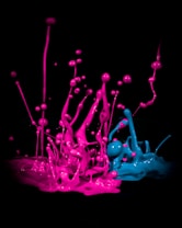 Vibrant splashes of neon pink and blue paint against a dark background, creating dynamic shapes and forms that appear to be mid-motion. The splashes rise in various directions with a sense of energy and chaos.