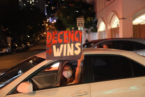 A person wearing a face mask is sitting inside a white car, holding up a handmade sign through the open window. The sign reads 'Decency Wins' in bold red and white letters against a black background. The scene is set on a city street at night, with buildings and parked cars visible in the background.