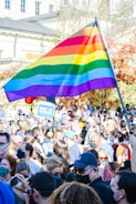 A large crowd of people gathered outdoors, many wearing face masks. A prominent rainbow flag is being held high above the group. Some individuals hold signs, including one that reads 'Biden Harris'. The background shows a building and trees with autumn foliage.