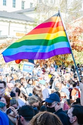 A large crowd of people gathered outdoors, many wearing face masks. A prominent rainbow flag is being held high above the group. Some individuals hold signs, including one that reads 'Biden Harris'. The background shows a building and trees with autumn foliage.