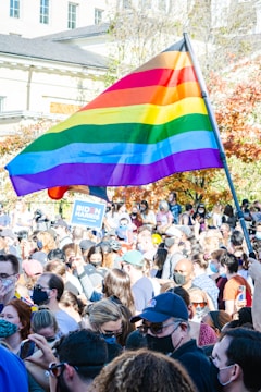 A large crowd of people gathered outdoors, many wearing face masks. A prominent rainbow flag is being held high above the group. Some individuals hold signs, including one that reads 'Biden Harris'. The background shows a building and trees with autumn foliage.
