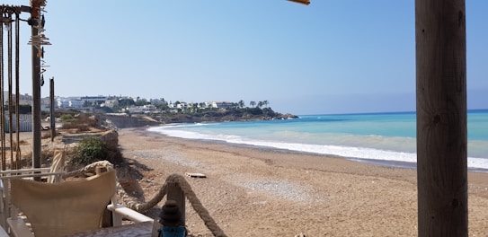 A sandy beach with gentle waves lapping against the shore, bordered by a rocky coastline with scattered shrubs. In the background, a small town with white buildings is visible, along with a row of tall palm trees atop a hill. A seaside cafe with rope barriers and wooden pillars is partially seen in the foreground.