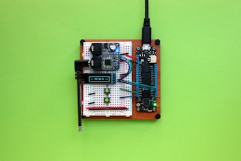 A breadboard setup with various electronic components including a microcontroller, jumper wires, resistors, and an OLED display showing a frequency. The components are mounted on a wooden base with a bright green background.
