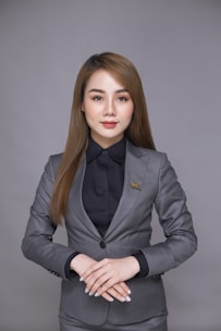 A person wearing a formal gray suit stands against a plain background. The suit includes a gray jacket over a black shirt, and there is a badge on the jacket. The hands are gently folded together at the front.